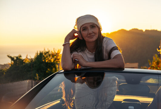 A smiling woman leans on a car during a summer sunset