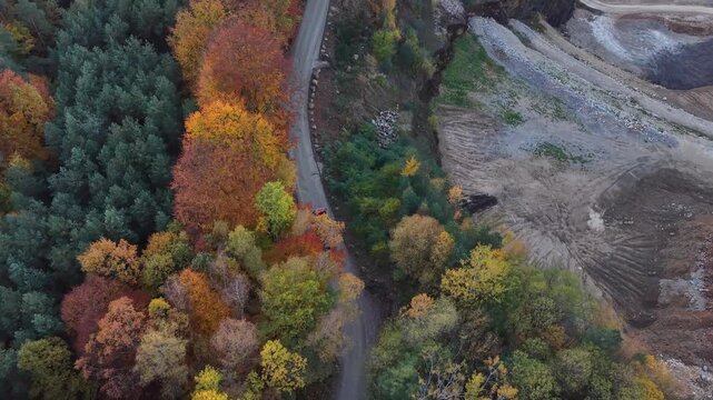 Dump truck driving along an autumn forest road toward a terraced industrial quarry. Aerial view of lorry riding though colorful trees, winding haul routes, and exposed earth. Revealing winding haul ro