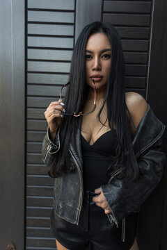 Cool urban lifestyle photo of a beautiful ethnic woman with long black hair in a leather jacket and top, posing gracefully near a dark textured wall.