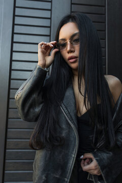 Cool urban lifestyle photo of a beautiful ethnic woman with long black hair adjusting her sunglasses, wearing a chic leather jacket near a dark wall. Pretty lady put on glasses