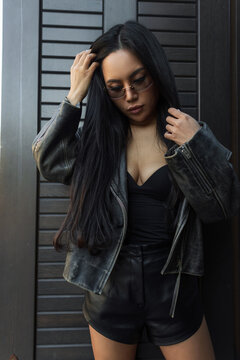 Stylish young woman touching long dark hair, wearing a black leather jacket, top, shorts, and trendy sunglasses, posing against a modern dark wall.