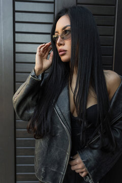 Stylish Asian woman with long dark hair wearing sunglasses and a leather jacket, looking away thoughtfully while posing against a modern dark wall.