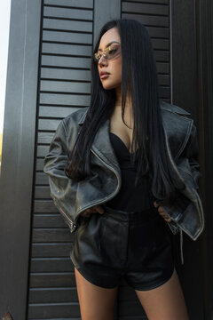 Stylish Asian woman with long dark hair wearing a leather jacket and sunglasses, looking to the side while posing against a dark slatted wall background.