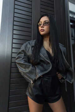 Stylish Asian woman in a leather jacket and sunglasses looking away confidently, posing in a medium shot against a dark slatted wooden wall background.