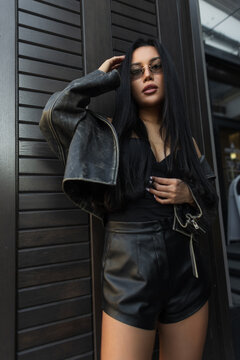 Low angle shot of a stylish Asian model in a dark leather jacket, black top, and shorts posing outdoors against a modern wooden panel wall. Pretty cool girl in black clothes on the street