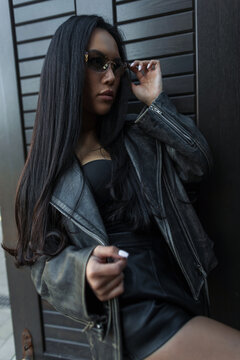 Medium portrait of a stylish Asian woman in a leather jacket and sunglasses touching her glasses, posing confidently outdoors against a dark slatted wall.