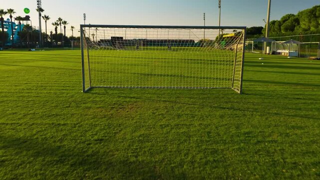 Aerial pullback away from a football goal across a green soccer field in Antalya, Turkey, with soccer goals, palm trees, manicured grass and resort recreation grounds near the Mediterranean coast