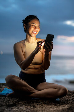 Asian woman taking a smartphone selfie while sitting in yoga pose on an ocean cliff