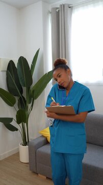 Professional female nurse talking on smartphone while taking notes on clipboard during domestic healthcare visit