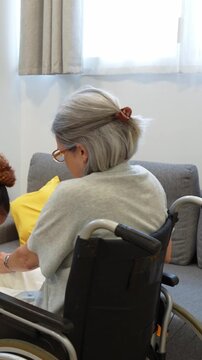 Dedicated latina home health nurse helping an elderly disabled woman standing up from a wheelchair to sit on sofa