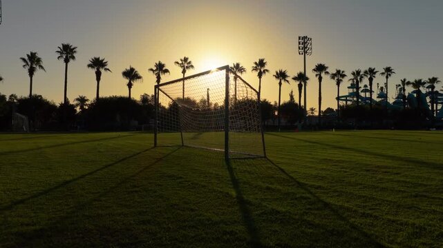 Forward aerial approach toward a football goal on a green field at sunrise in Antalya, Turkey, with soccer goals, palm trees, manicured grass and resort recreation grounds near the Mediterranean coast