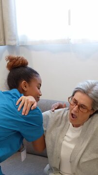 Caring latina home health nurse helping an elderly woman stand up from the sofa and walk slowly indoors during a routine visit