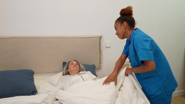 Caring latin home health nurse tucking in an elderly woman in bed and gently touching her cheek during a medical visit