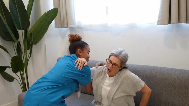 Compassionate latina caregiver assisting an elderly woman with back pain to stand up from the sofa and walk at home