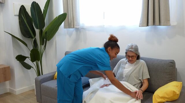 Caring latina home health nurse covering an elderly woman with a white blanket and giving her a pillow on the sofa at home