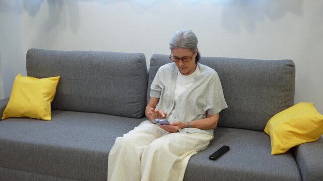 Senior woman sitting on couch, holding smartphone and talking during home healthcare visit from female nurse