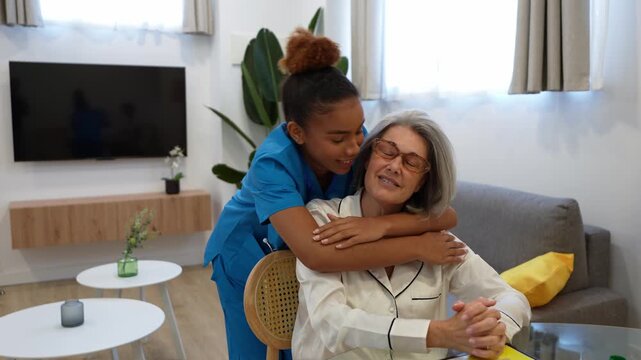 Cheerful latina home health nurse hugging an elderly woman seated at the table, showing affection and care during a home visit