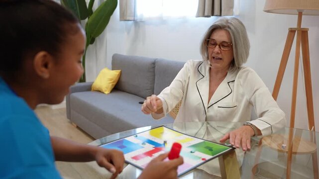 Young latina nurse playing a board game with an elderly woman in her living room, enjoying recreational cognitive therapy at home