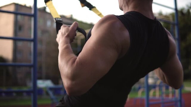 Muscular man performing inverted row exercise with suspension straps at an outdoor gym for strength and endurance training