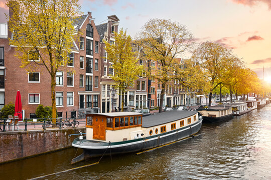 Amsterdam, Netherlands. Vintage brick canal houses line the waterfront at sunset. Classic European architecture with traditional houseboats and autumn trees captures a scenic retro city vibe.