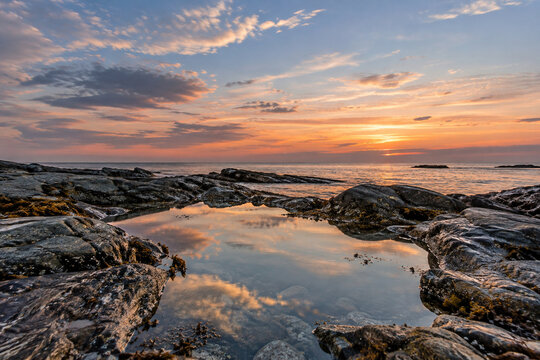 Calm tide pool among dark rocks on the seashore reflecting soft sunset clouds