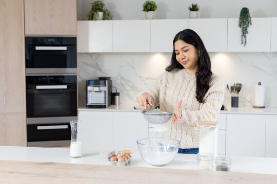 Asian woman holding fine mesh sieve while sifting flour into glass bowl at island