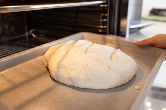 Hand holding metal baking tray with round bread dough near open oven and flour specks