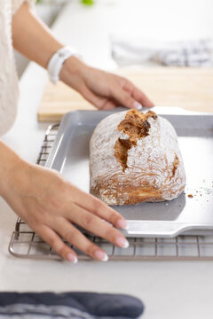 Hands with white wristwatch hovering near metal tray holding rustic loaf on cooling rack