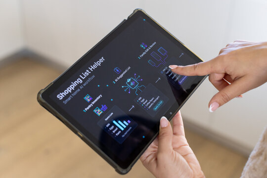 Woman holding tablet in rugged black case while touching Shopping List Helper screen near pale wall