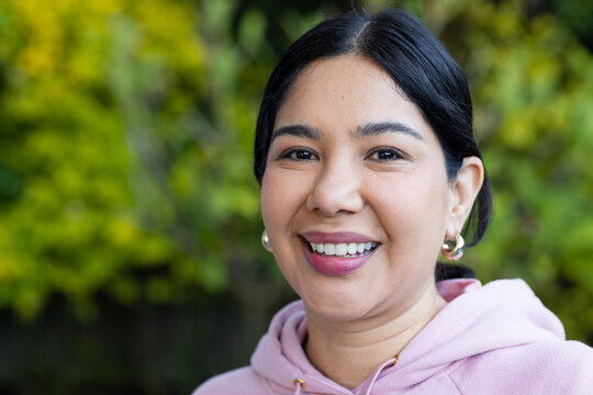 Asian woman in pink hoodie and hoop earrings smiling at camera against blurred foliage, copy space