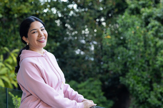 Woman in light pink hoodie standing beside glass railing looking toward camera amid trees