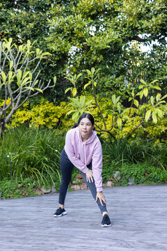 Asian woman in pink hoodie and black leggings stretching on paved walkway near green shrubs