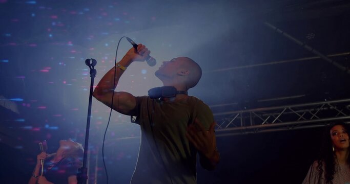 Bald vocalist singing into handheld microphone on stage under colored lights amid haze