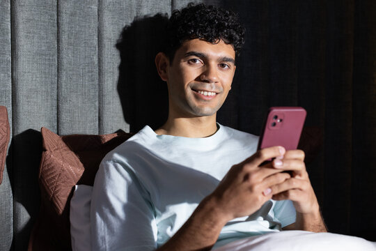 African American man with dark curly hair leaning on brown cushions and holding pink smartphone