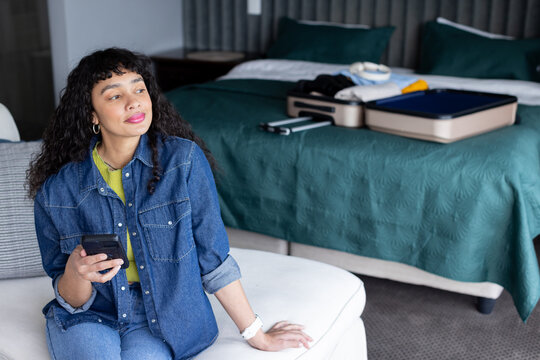 African American woman sitting on cushioned seat holding smartphone and looking away in hotel room