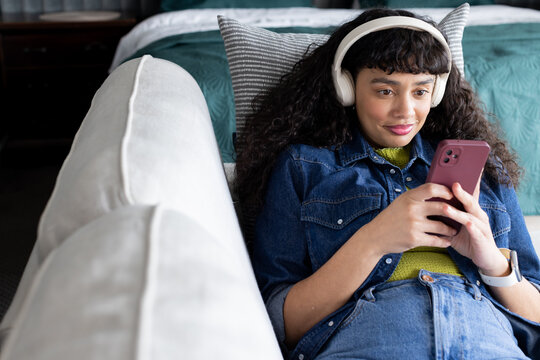 African American woman wearing headphones reclining on white sofa while holding burgundy smartphone