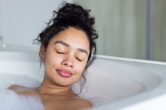 African American woman resting in white bathtub filled with foam, eyes closed, tilting face up