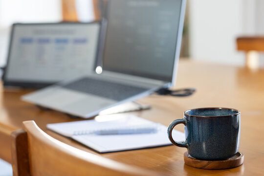 Wooden table holding two open laptops, notepad, pen and speckled ceramic mug near wooden chair back