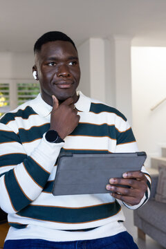 African American man sitting in bright room holding tablet and touching chin wearing sweater