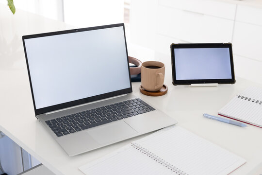White desk holding silver laptop, tablet, beige mug and notebooks near plant leaves, copy space