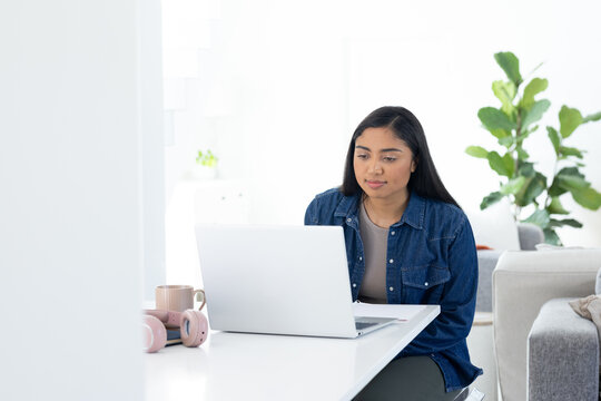 African American woman sitting at white table using silver laptop near pink headphones and pale mug