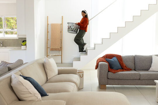 African American woman carrying gray laundry basket while stepping up stairs in modern home
