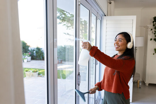 African American woman spraying glass patio door while holding blue squeegee in bright home