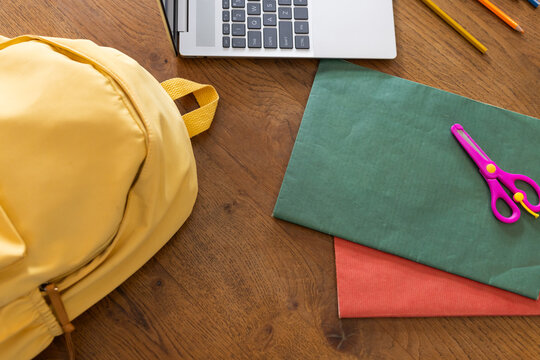 Yellow backpack, open silver laptop and green paper lying beside purple scissors on wooden tabletop