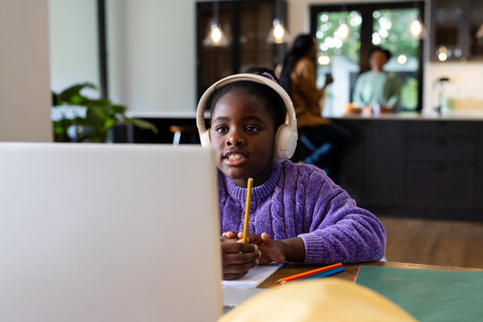 African American teenage girl wearing headphones holding yellow pencil while using laptop at table