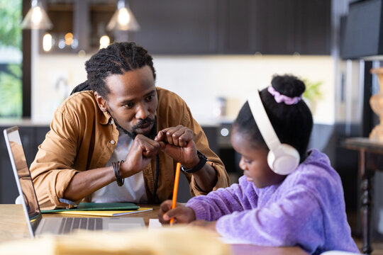 African American man and girl sitting at table, girl writing in notebook near open laptop