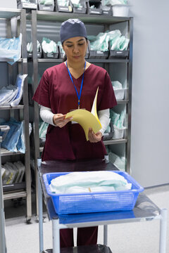 Asian woman in maroon scrubs and blue cap inspecting yellow package in clinical supply room