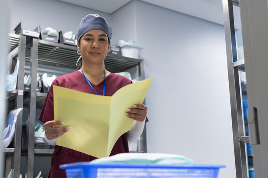 Woman in maroon scrubs and blue surgical cap holding open yellow folder in clinical supply room