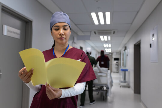 Indian woman in scrubs reading yellow folder in corridor while workers standing near gurney