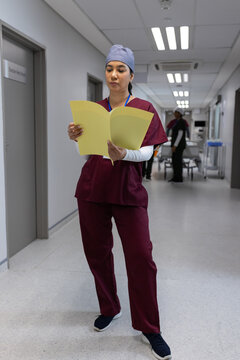 Indian woman in maroon scrubs reading open yellow folder while walking in hospital hallway